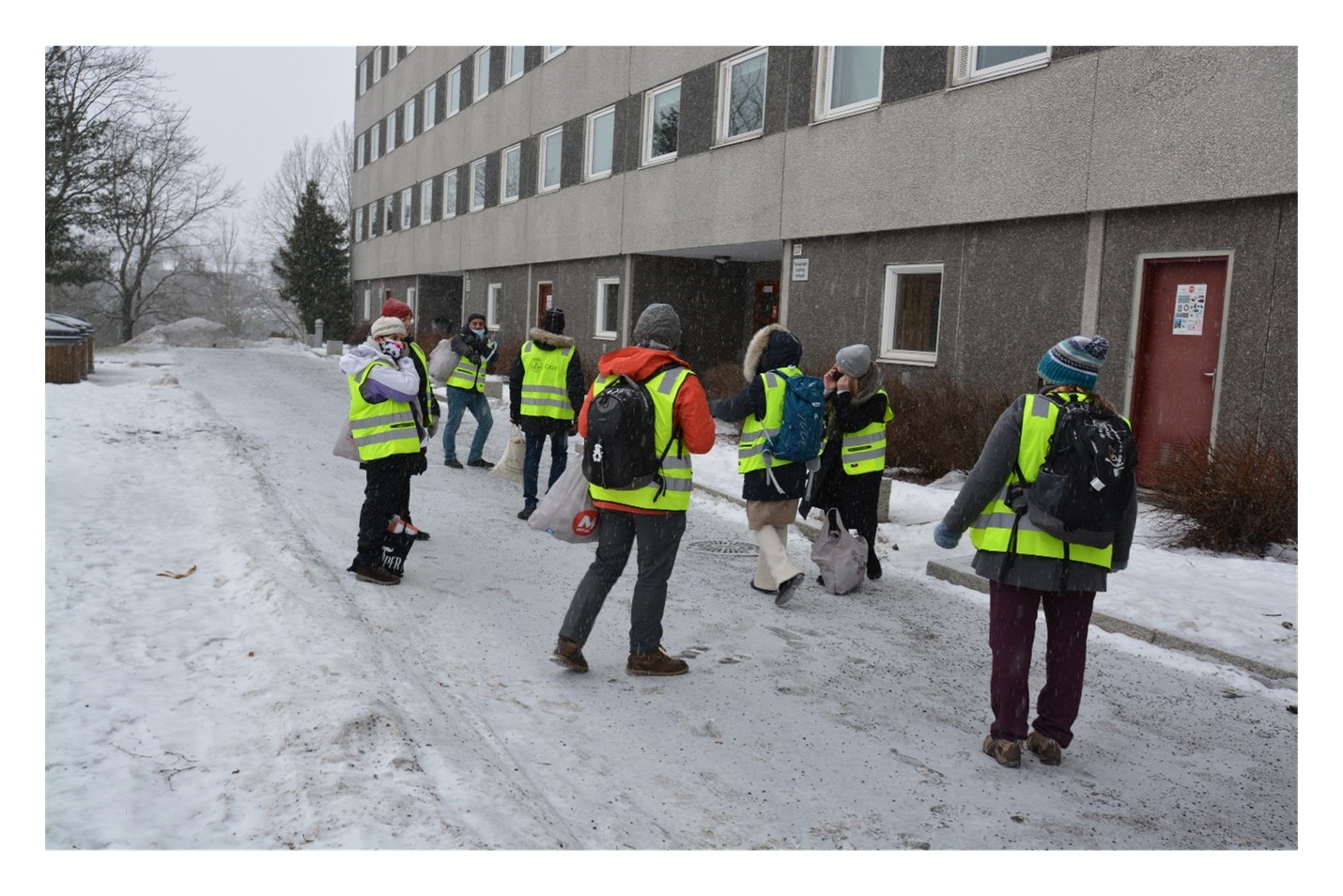 Dør til dør aksjon med personer iført refleksvest og munnbind utenfor en blokk 